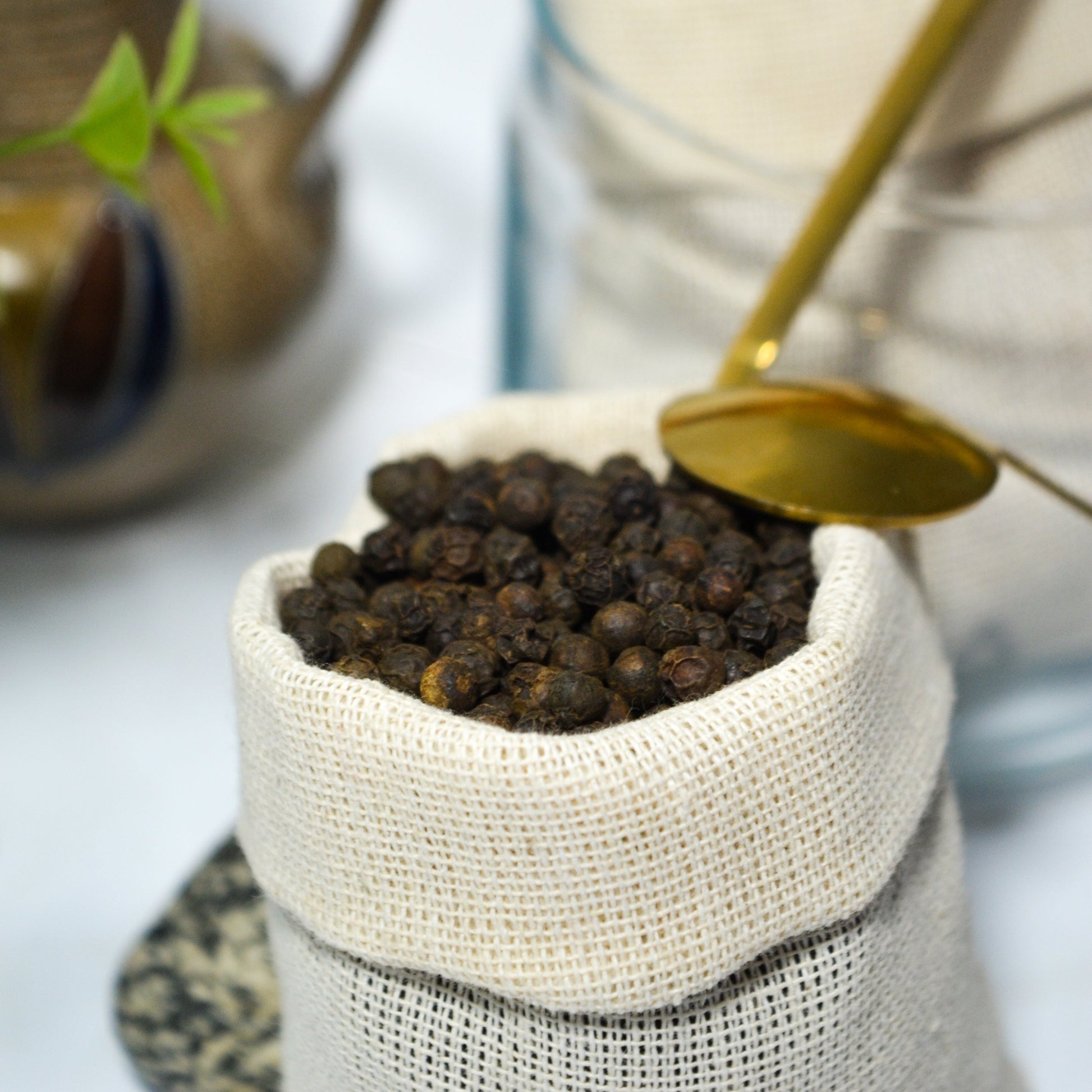 Black peppercorns in a woven container with a spoon, cinnamon sticks, and a teapot in the background.