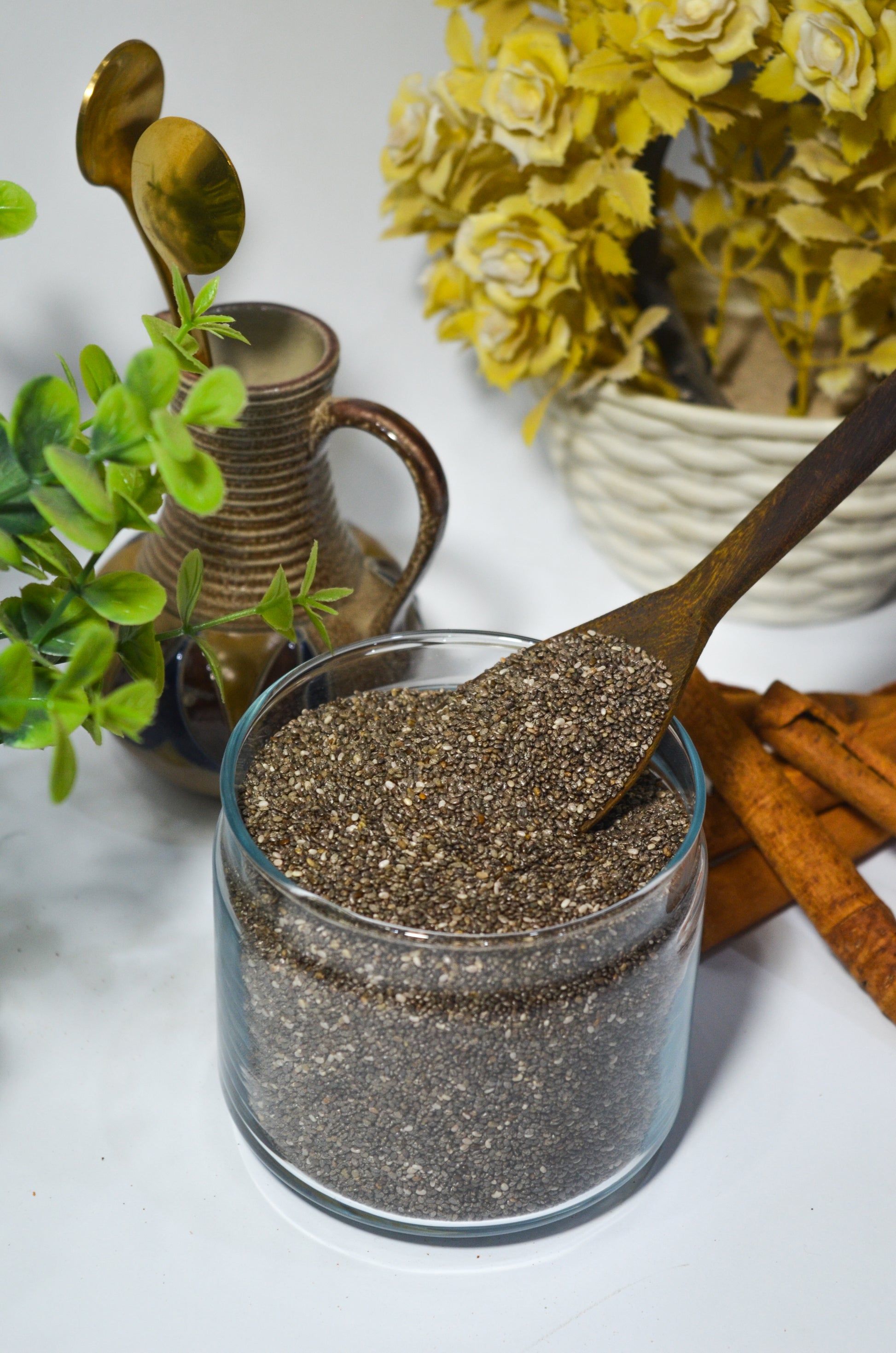 Glass jar of chia seeds with a wooden spoon, surrounded by plants and cinnamon sticks on a white surface.