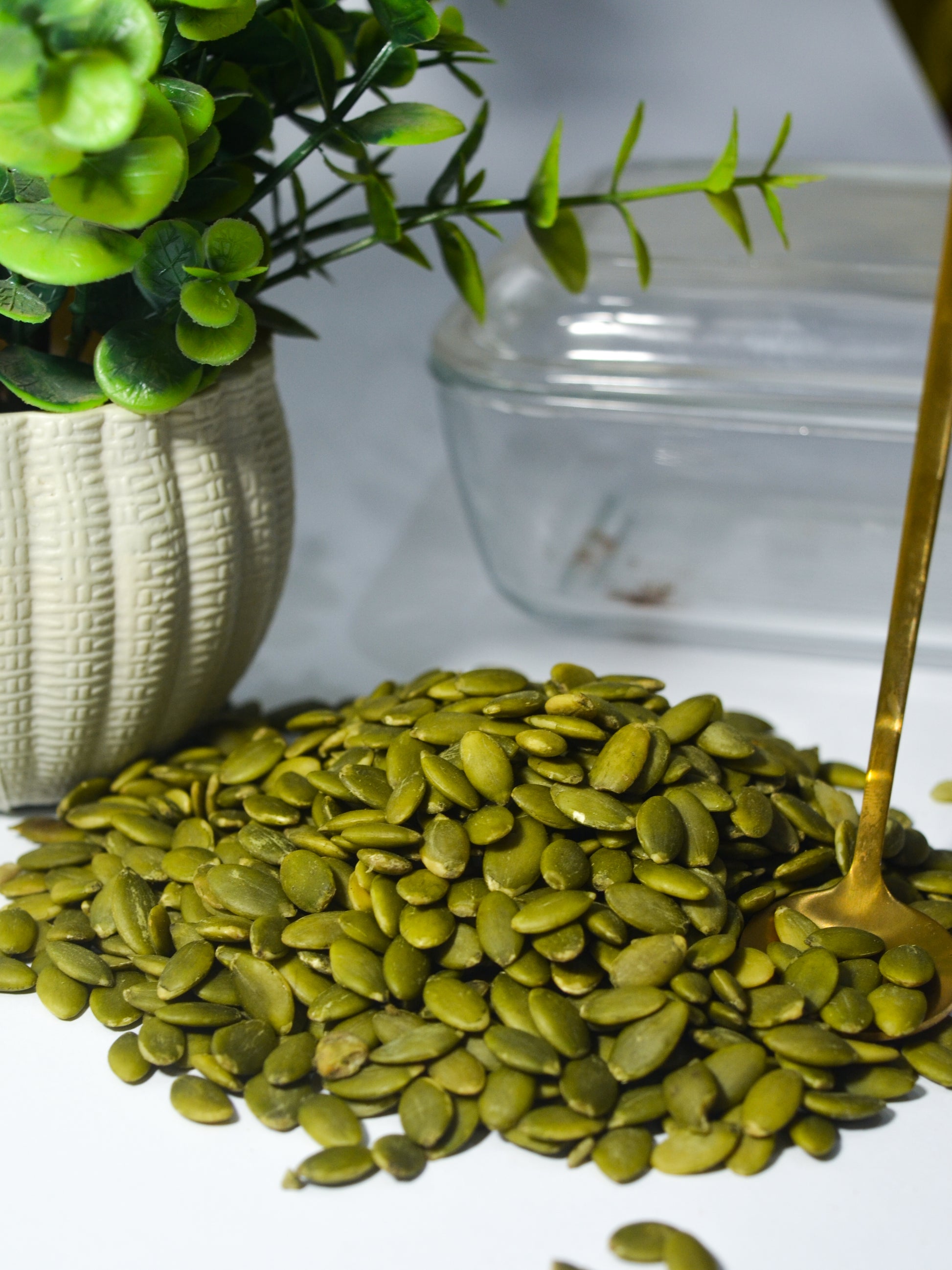 Pile of green pumpkin seeds with a small plant and glass container in the background.